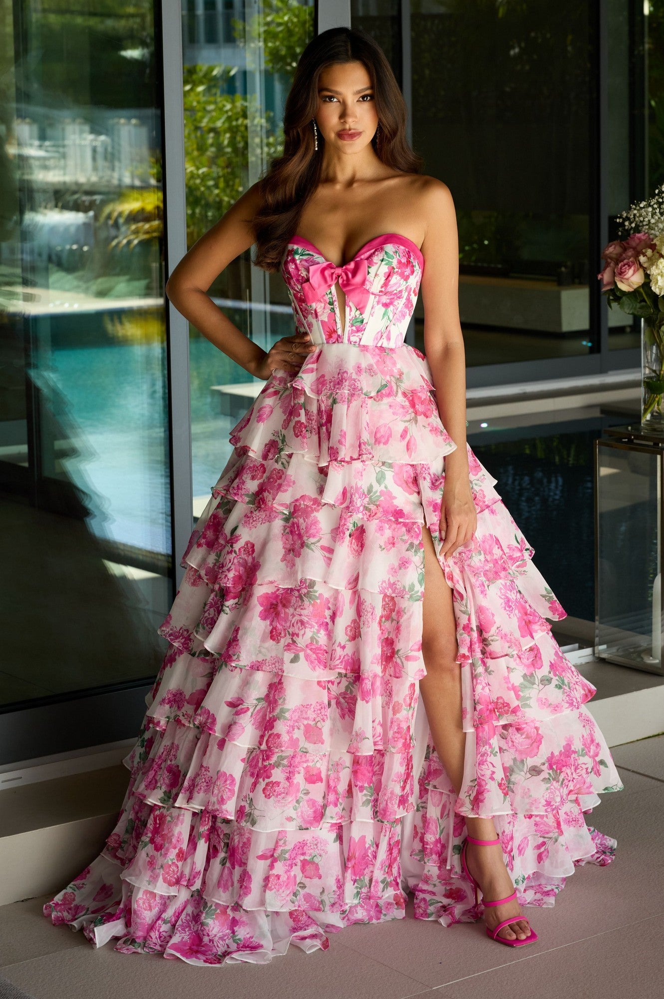 A woman stands indoors by a window wearing the Strapless Long Print Prom Dress: Ava Presley 42207, featuring floral print, ruffles, a high slit, and matching heels.